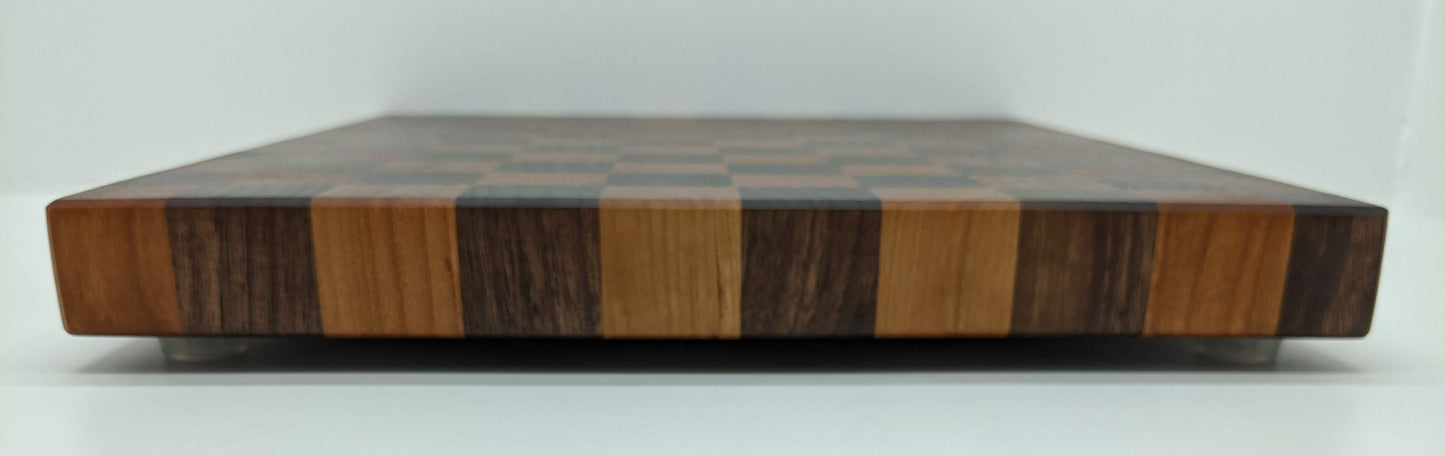 A full, side view of a handcrafted Walnut and Cherry end‑grain cutting board featuring a checkered pattern of alternating dark and light hardwood blocks. The board is shown elevated on rubber feet, highlighting its stability, smooth finish, and premium craftsmanship—ideal for chef‑quality prep, charcuterie, gifting, and high‑end kitchen décor.