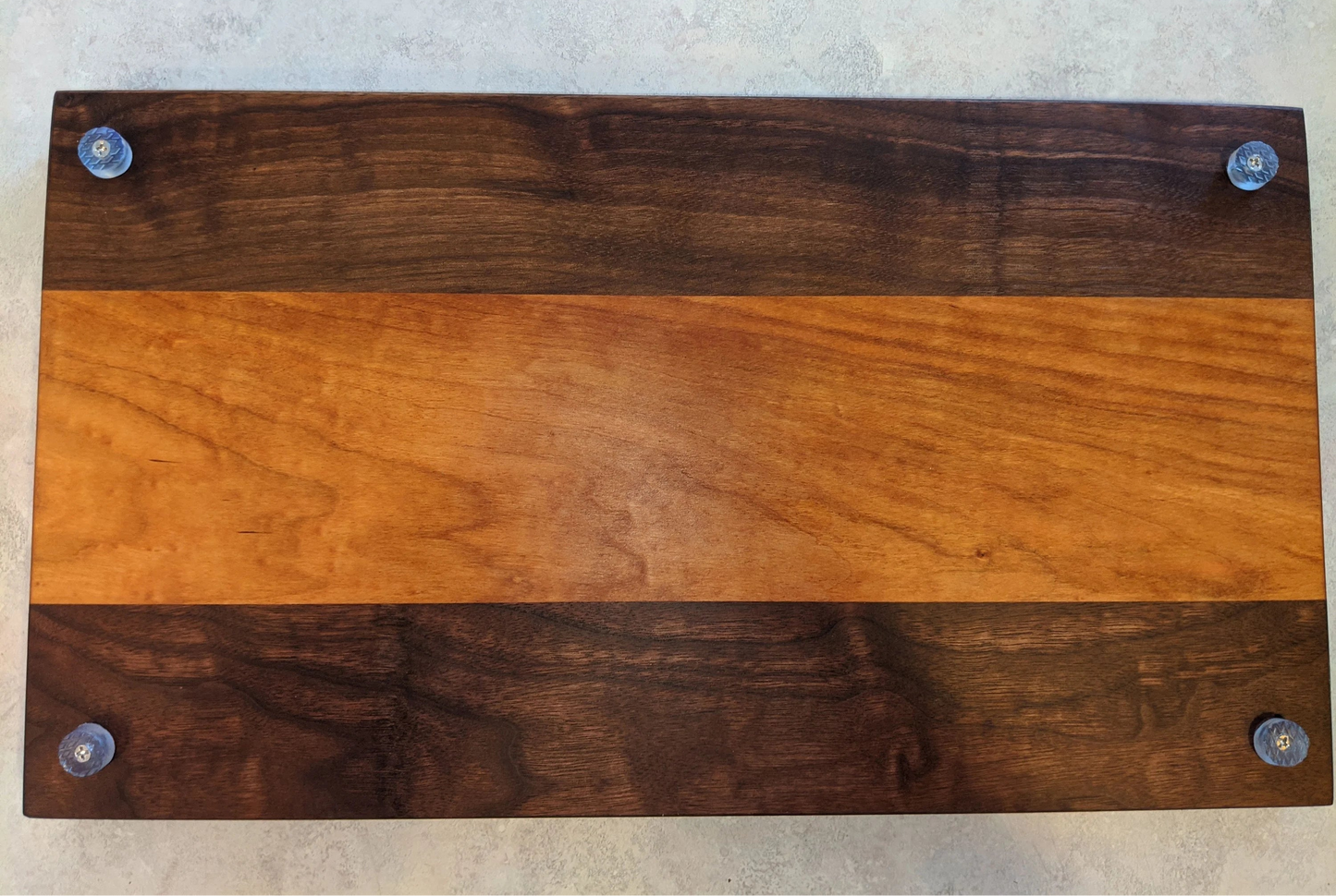 A handcrafted Walnut and Cherry cutting board with a warm Cherry center strip and dark Walnut outer panels, shown from the bottom with four non‑slip rubber feet attached at each corner. The smooth, polished hardwood surface and contrasting grain patterns highlight premium craftsmanship — ideal for shoppers searching for a Walnut and Cherry cutting board, handmade kitchen gift, or artisan charcuterie board.