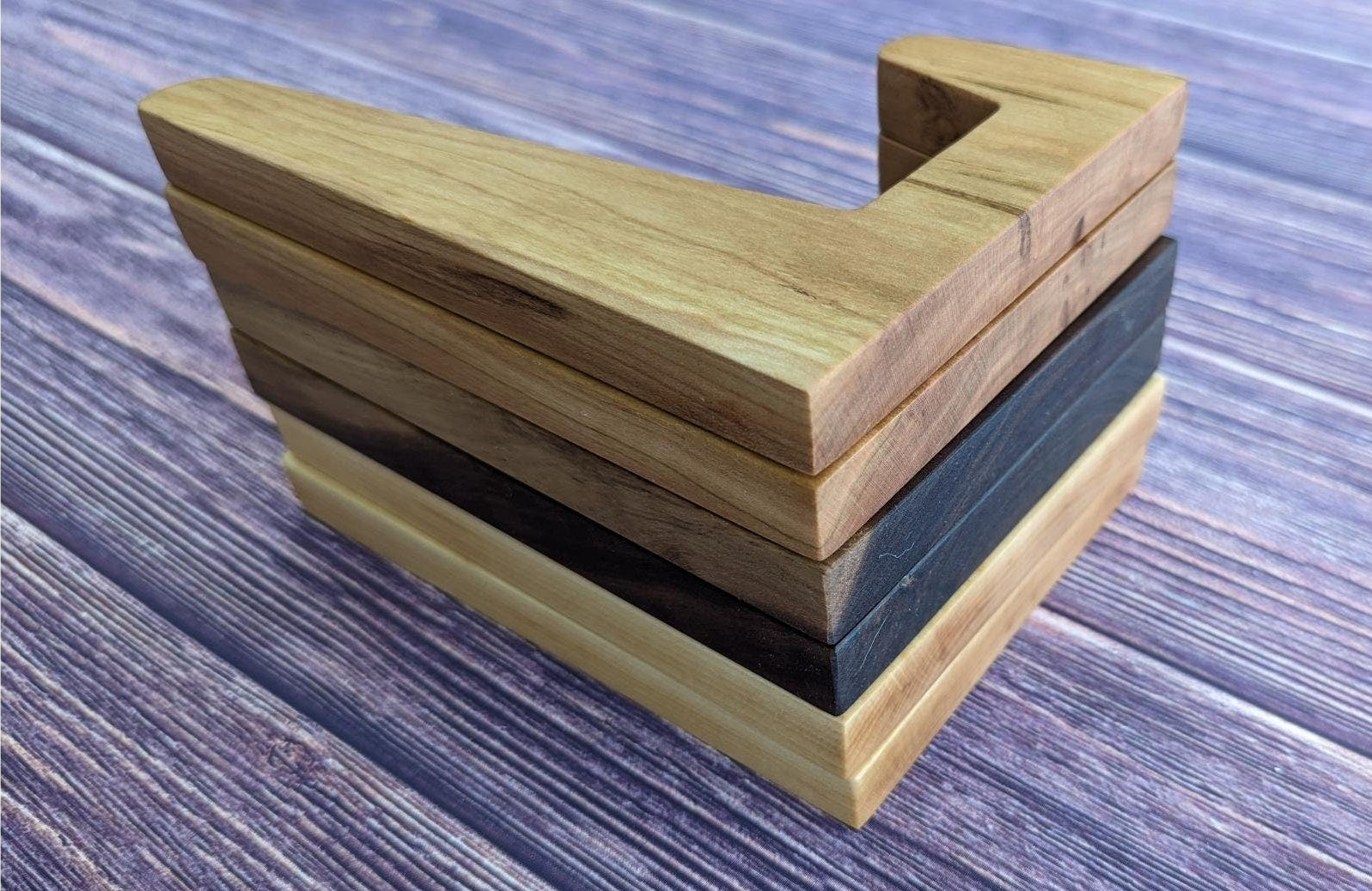 A stack of L-shaped wooden cutting board stands in varying wood species, including Maple, Cherry, and Walnut. The vertical stack shows the consistent 90-degree profile and smooth, rounded edges of each holder. This close-up highlights the natural wood grain and high-quality of the pieces. Handcrafted by Great Scott Woodworks, these versatile stands are designed for organized kitchen storage, displaying heavy boards, or holding tablets and cookbooks.