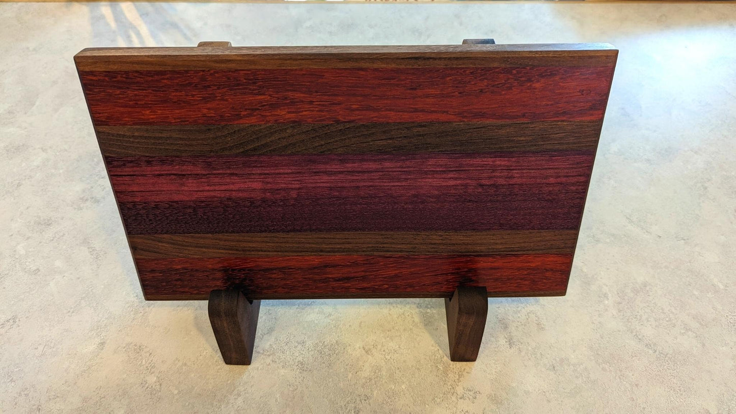 A long, multi-colored hardwood cutting board with deep purple and red grain displayed horizontally on a pair of walnut L-shaped stands. The side view highlights the 90-degree support and how the stands securely hold a thick, heavy board. These handcrafted wooden holders from Great Scott Woodworks provide a stable, space-saving solution for kitchen countertops, allowing for both beautiful display and efficient air-drying after use.