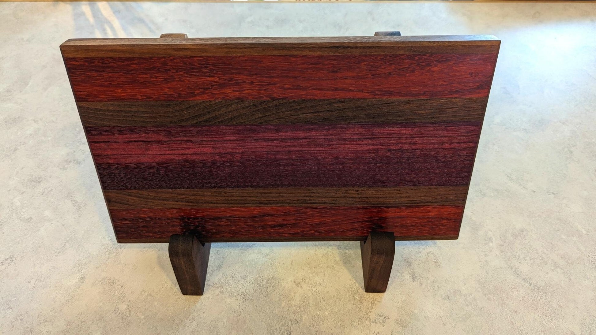 A long, multi-colored hardwood cutting board with deep purple and red grain displayed horizontally on a pair of walnut L-shaped stands. The side view highlights the 90-degree support and how the stands securely hold a thick, heavy board. These handcrafted wooden holders from Great Scott Woodworks provide a stable, space-saving solution for kitchen countertops, allowing for both beautiful display and efficient air-drying after use.