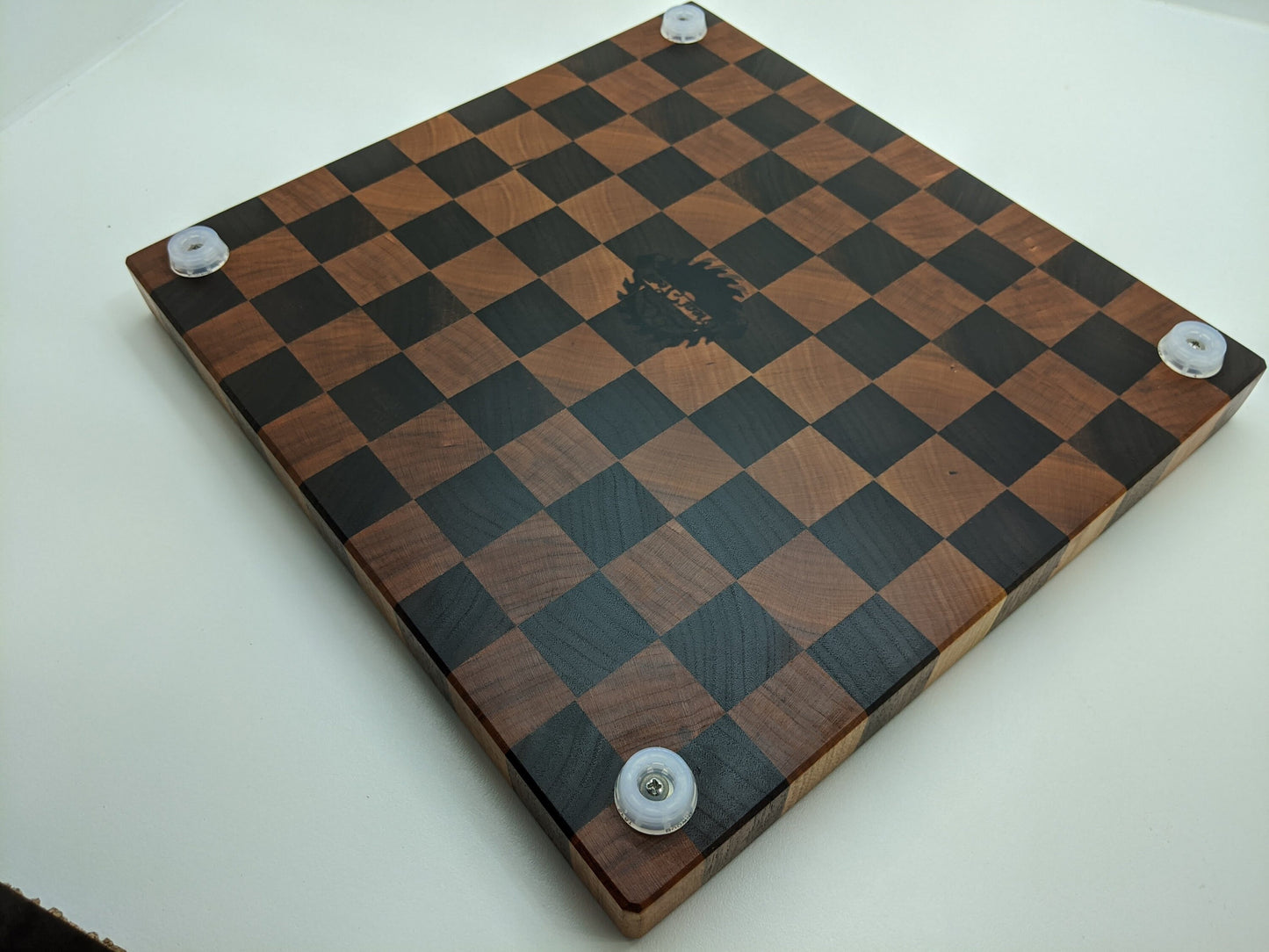 The underside of a handcrafted Walnut and Cherry end‑grain cutting board featuring four sturdy rubber feet for stability and countertop protection. The image also shows this shops logo etched in the center, along with the alternating dark and light hardwood blocks and smooth finish, highlighting premium craftsmanship for chef‑quality kitchen use.