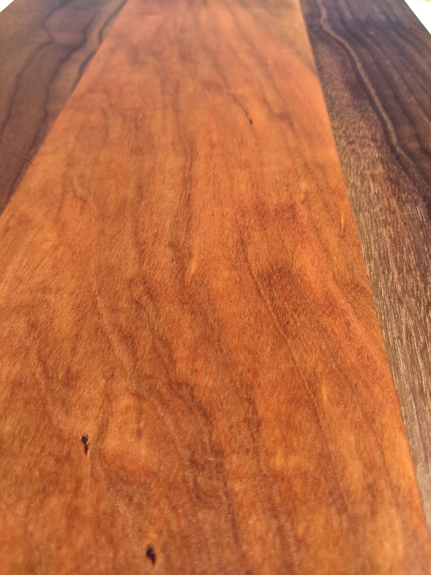 A close‑up of a handcrafted Walnut and Cherry cutting board featuring a warm Cherry center panel framed by two dark Walnut outer strips. The smooth, polished surface and visible grain patterns highlight the board’s premium craftsmanship — ideal for shoppers searching for a Walnut and Cherry cutting board, handmade kitchen gift, or artisan charcuterie board.