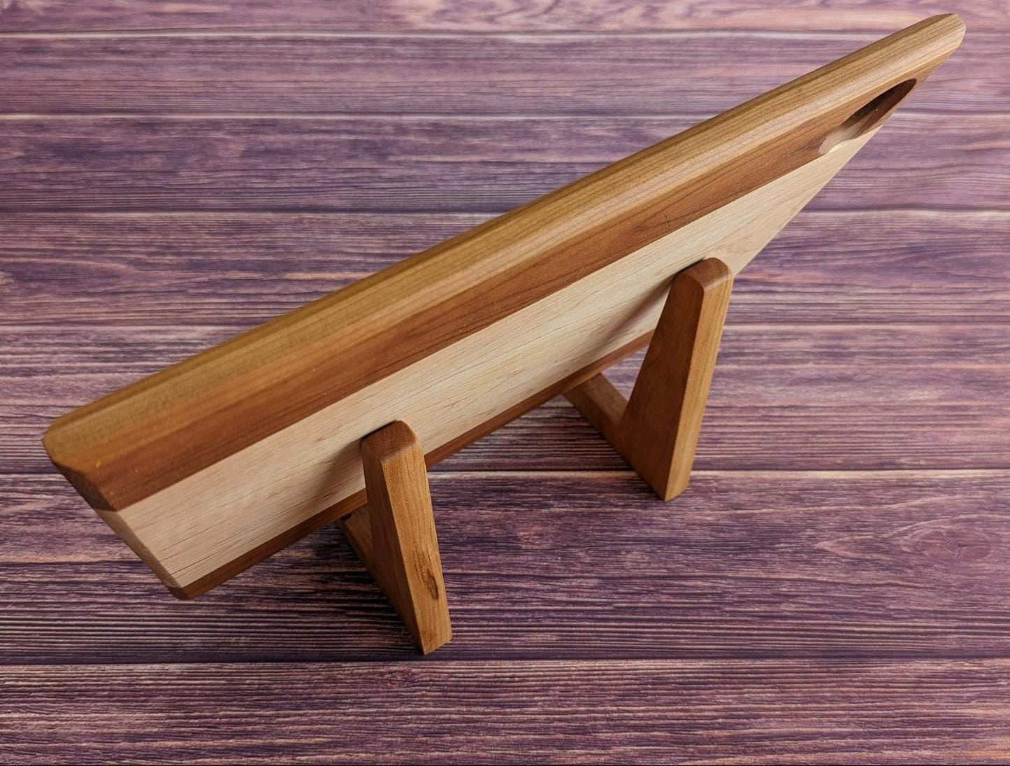 Rear view of a large handcrafted cutting board supported by two maple L-shaped stands on a purple wood surface. This angle highlights the 90-degree upright support and how the stands discreetly hold heavy boards for display or drying without cluttering the counter. Made by Great Scott Woodworks, these solid wood holders are a sturdy and minimalist solution for kitchen organization, charcuterie board storage, or air-drying after washing.