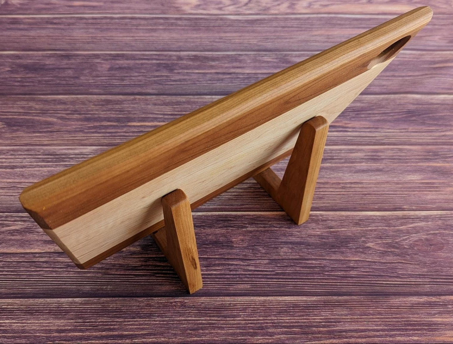 Rear view of a large handcrafted cutting board supported by two maple L-shaped stands on a purple wood surface. This angle highlights the 90-degree upright support and how the stands discreetly hold heavy boards for display or drying without cluttering the counter. Made by Great Scott Woodworks, these solid wood holders are a sturdy and minimalist solution for kitchen organization, charcuterie board storage, or air-drying after washing.