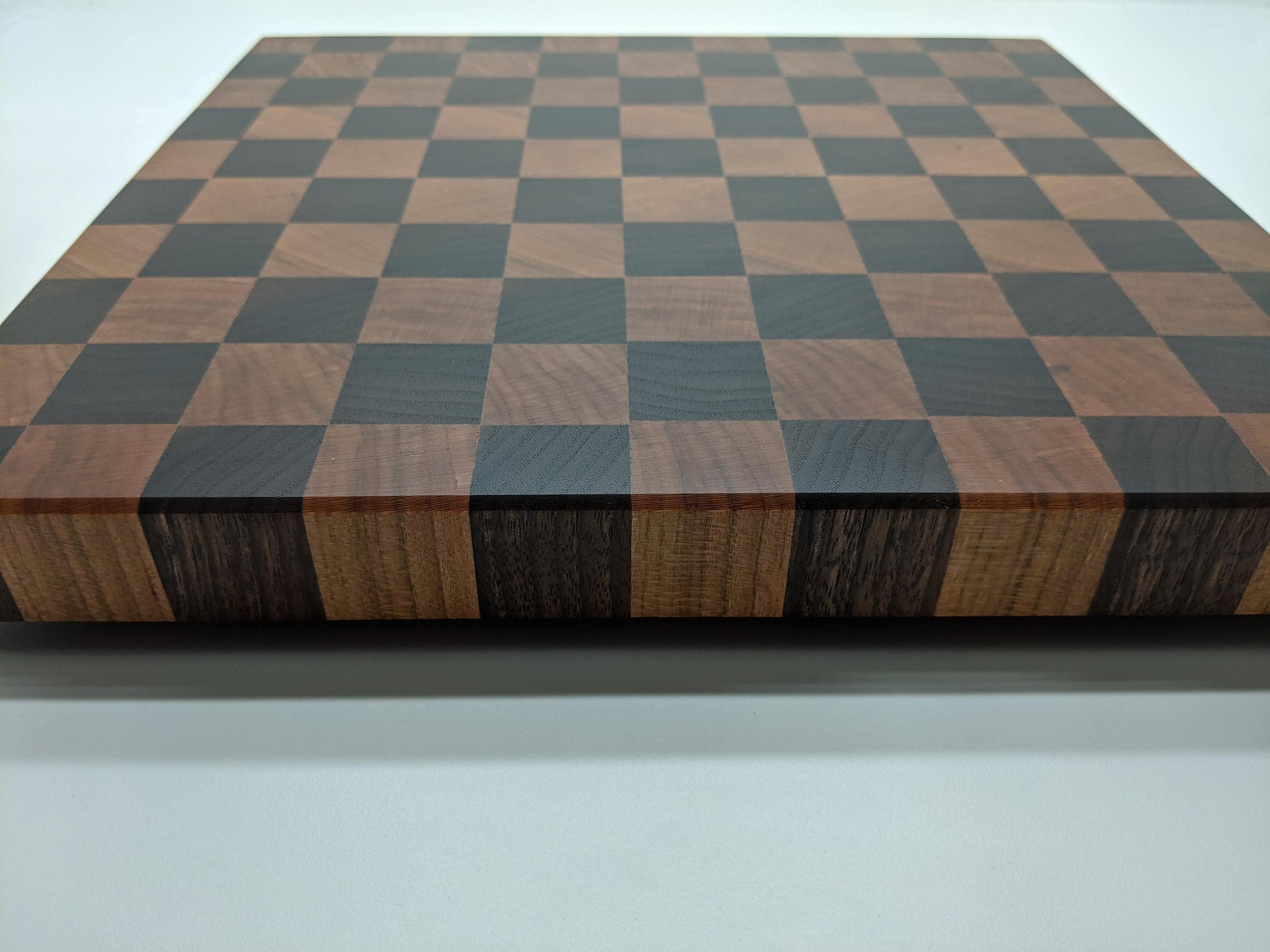 A handcrafted Walnut and Cherry end‑grain cutting board shown at an angled view, highlighting how the alternating dark and light hardwood blocks wrap seamlessly over the edges. The checkered pattern, smooth finish, and precise joinery showcase premium craftsmanship, ideal for chef‑quality prep, charcuterie, gifting, and high‑end kitchen décor.