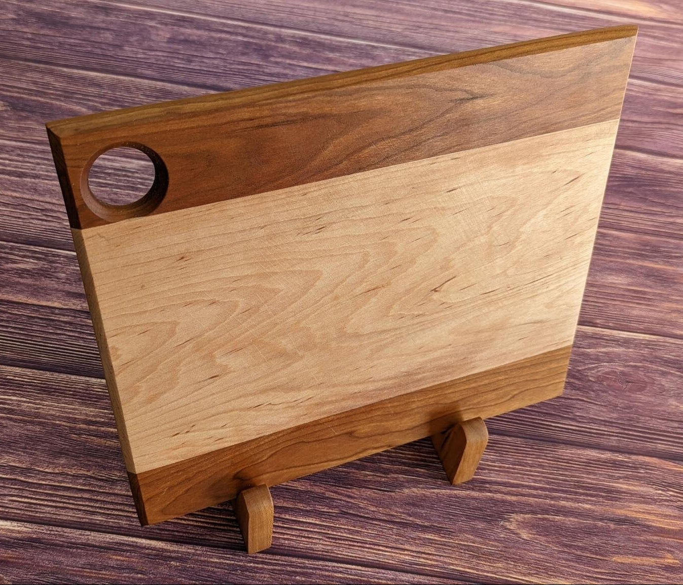 Front-facing view of a thick maple cutting board with walnut accents held vertically by two small L-shaped wooden stands. The board features a circular finger hole in the corner and sits securely in the 90-degree supports on a dark, rustic wood surface. Handcrafted by Great Scott Woodworks, these stands provide a stable, space-saving way to display heavy butcher blocks or air-dry boards after washing without taking up excess kitchen counter space.